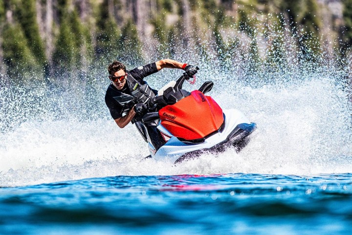 Person riding a red and white jet ski, creating spray, with a forested background.