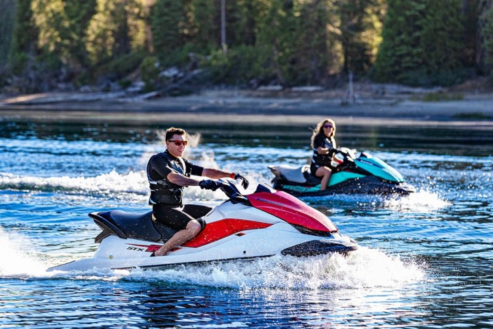 Two people riding jet skis on a calm lake with forested shoreline.