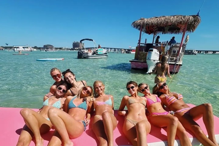 Group of people in swimsuits on a float in clear water with boats and blue sky in the background.