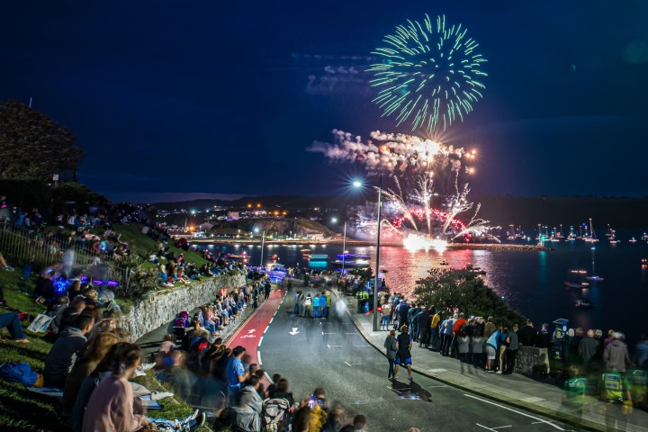 Crowd watching fireworks over a harbor at night, with a road and grassy area in the foreground.