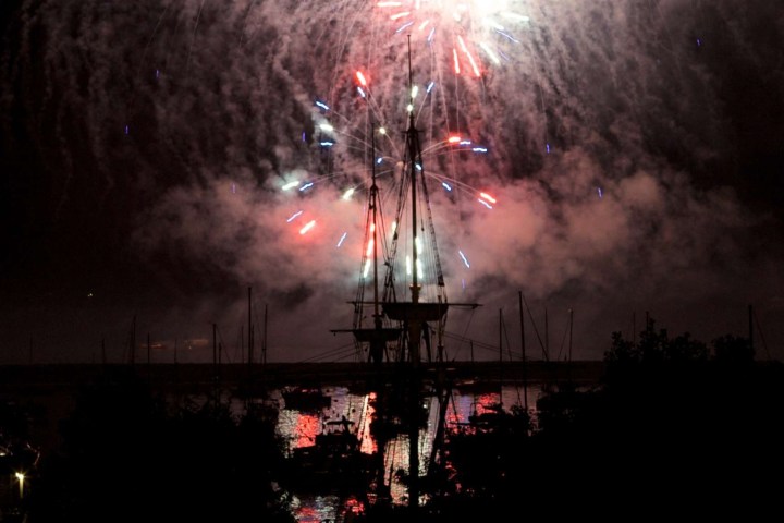 Silhouette of a ship under fireworks in a night sky over water.