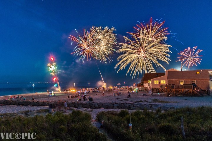 Fireworks display over a beach with people watching and houses nearby, under a clear night sky.