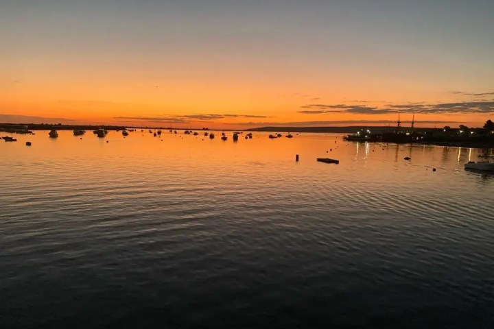 Sunset over calm water with boats silhouetted and an orange sky.